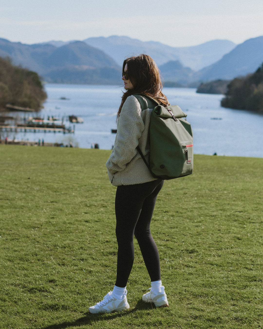 Female model walking in the hills of the lake district wearing our stylish, multi-functional moss green roll-top rucksacks, which have extra storage and waterproof outer for all-weather protection. Perfect as an after swim coat, surf robe, beach coat and dog walking jacket. 