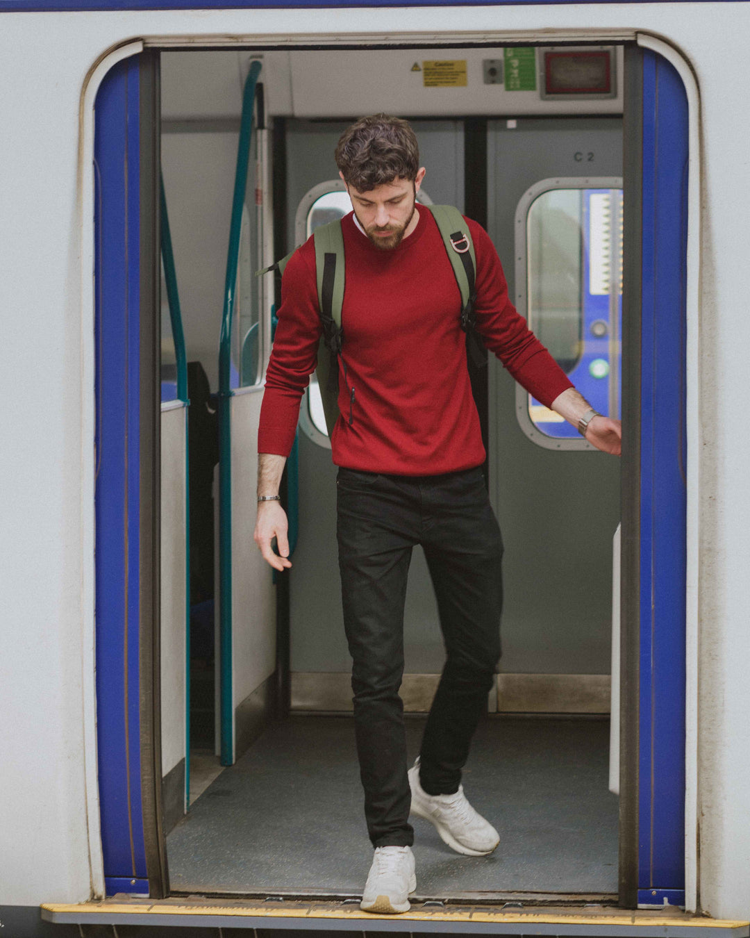 Model commuting on the train wearing our unisex extra-fine merino wool 1/4 zip jumper in maroon red, made from a lightweight fabric and reactive natural fibre. Ideal for layering under our waterproof dry robes for spectating touchdown sports or walking the dog. The perfect addition to any minimalist capsule wardrobe.