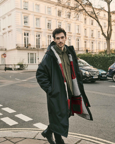 A man wearing a functional and stylish, oversized dry robe around the city in the colour anthracite. The hooded changing robes for men and women are made from recycled materials, with fleece-lined, waterproof pockets, adjustable hood and cuffs, and velcro straps for a personalised fit.