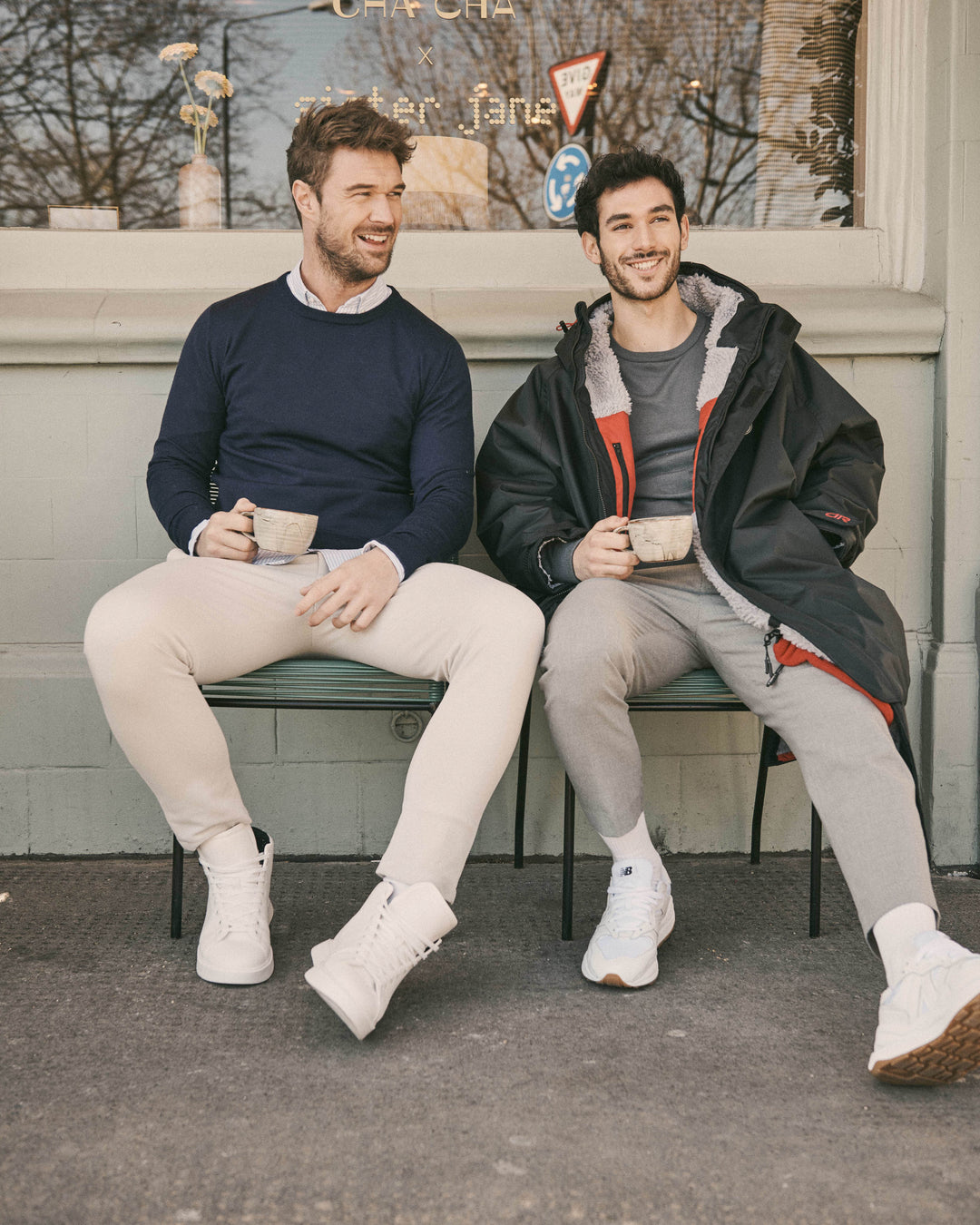 Two men sitting in a cafe in London drinking coffee wearing extra-fine merino wool knit jumpers with a fleece-lined, waterproof and weatherproof outdoor jacket that can be worn as an after swim coat, dog walking jacket, rugby coat, athletic coat or football coat.