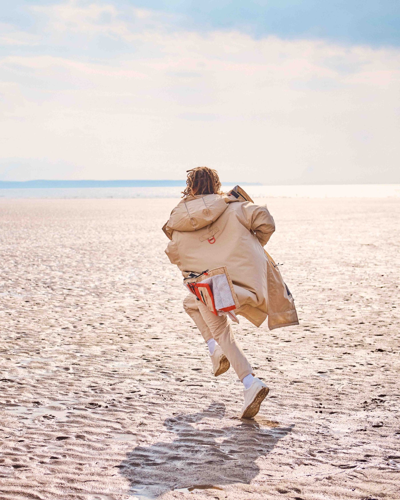 A Child running on a beach wearing a beige dry-robe by D-Robe. These hooded unisex changing robes are made from recycled materials, with a waterproof outer, fleece-lined inner, waterproof pockets, adjustable hood and cuffs. It is perfect for a stroll on the beach and guarantees to keep you warm and dry. 