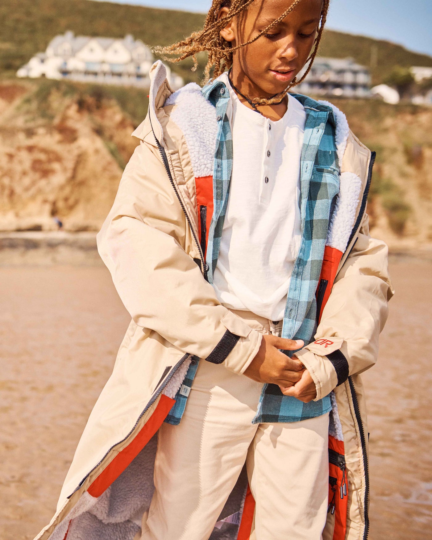 A Child standing on a beach wearing a beige dry-robe by D-Robe over his checked shirt and pants. These hooded unisex changing robes are made from recycled materials, with a waterproof outer, fleece-lined inner, waterproof pockets, adjustable hood and cuffs. It is perfect for a stroll on the beach and guarantees to keep you warm and dry. 