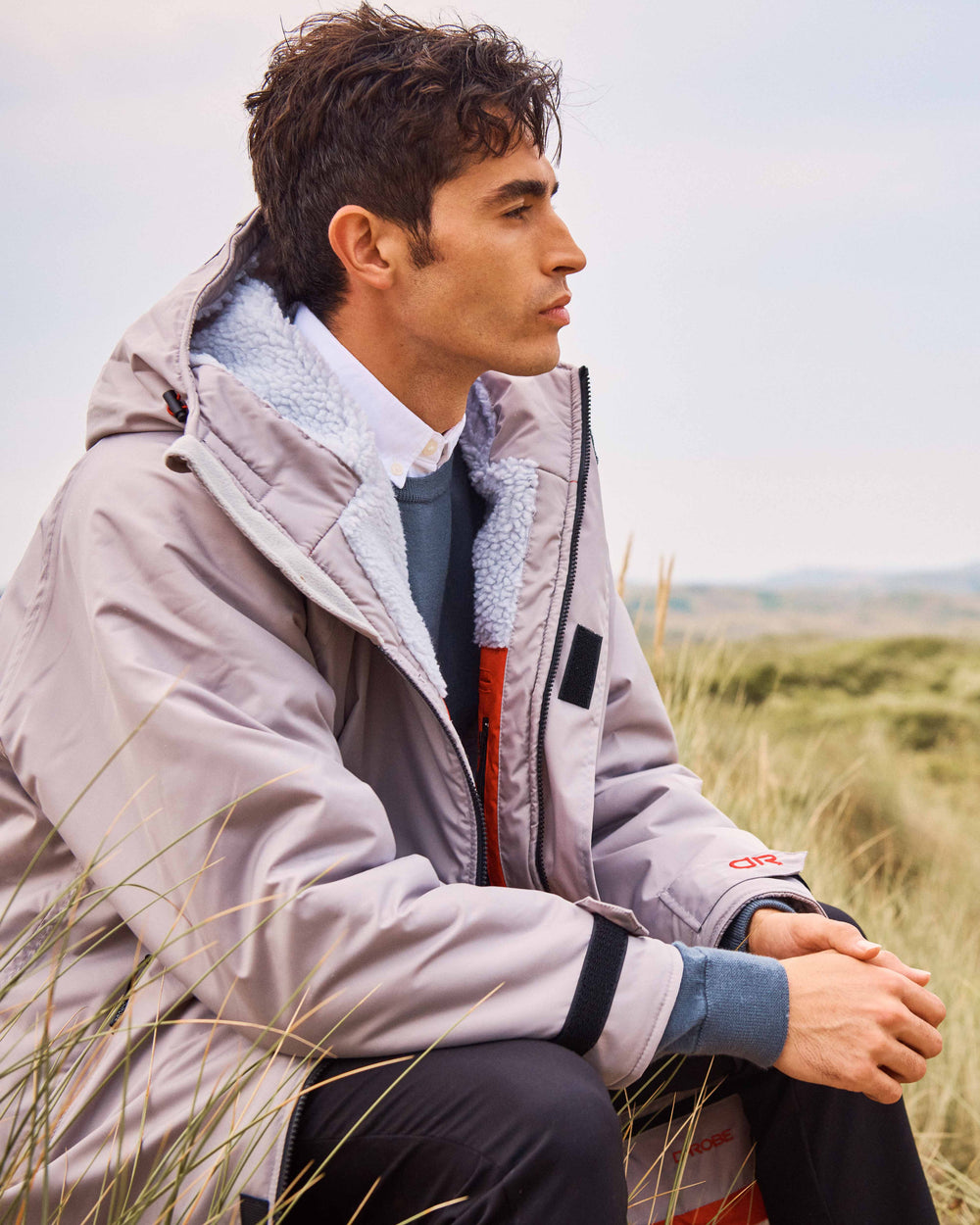 There is a man seated down wearing a grey dry-robe by D-Robe and black plants at Devon Beach. These hooded unisex changing robes are made from recycled materials, with a waterproof outer, fleece-lined inner, waterproof pockets, adjustable hood and cuffs. It is perfect for a stroll on the beach on a Spring Day.
