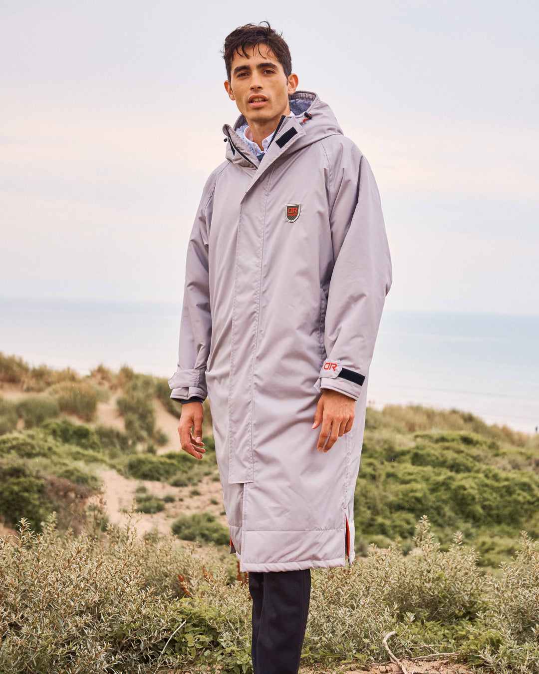  A man wearing a zipped-up dry-robe by D-Robe at Devon Beach. This robe features tapered seams to make it waterproof. These hooded unisex changing robes are made from recycled materials, with a waterproof outer, fleece-lined inner, waterproof pockets, adjustable hood and cuffs. It is perfect for a stroll on the beach on a Spring day.
