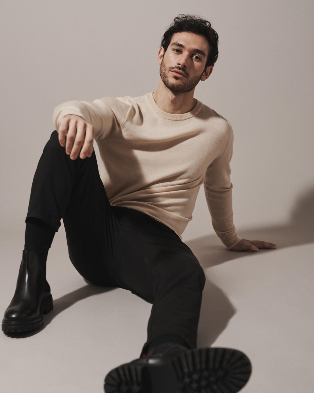 Model wearing our unisex extra-fine merino wool crew neck jumper in desert sand, made from a lightweight fabric and reactive natural fibre. Ideal for layering under our waterproof dry robes for smart city commute attire. The perfect addition to any minimalist capsule wardrobe.