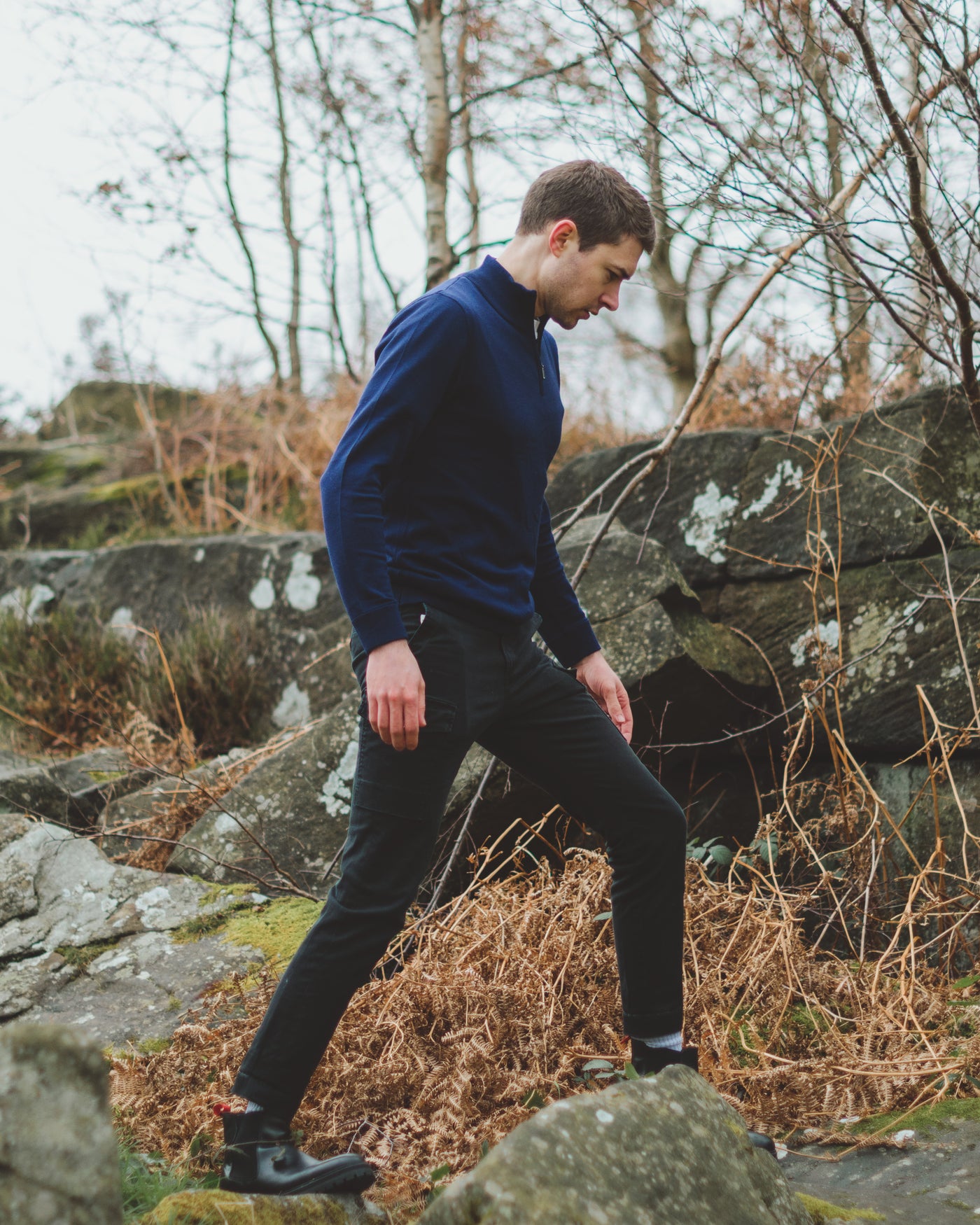 Model walking in the woods wearing our unisex extra-fine merino wool 1/4 zip jumper in navy, made from a lightweight fabric and reactive natural fibre. Ideal for layering under our waterproof dry robes for spectating touchdown sports or walking the dog. The perfect addition to any minimalist capsule wardrobe.